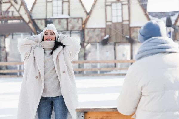 Sorrindo Jovem Mulher Ajustando Chapéu Inverno Perto Namorado Borrado — Fotografia de Stock