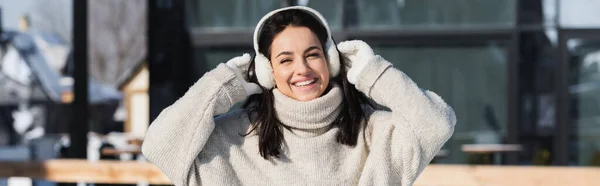 cheerful young woman in sweater and ear muffs in wintertime, banner