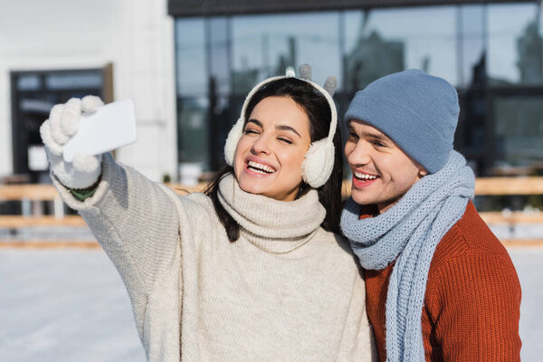 happy woman in sweater and ear muffs taking selfie with cheerful boyfriend on ice rink 
