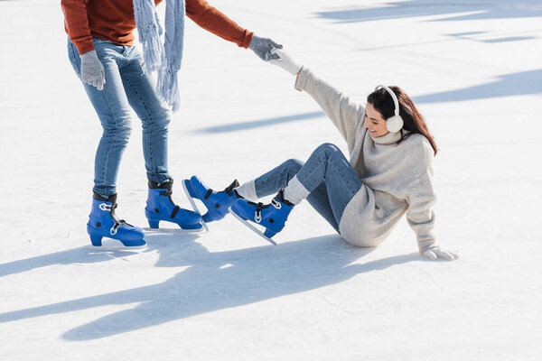 man on ice skates helping smiling girlfriend get up on ice rink 