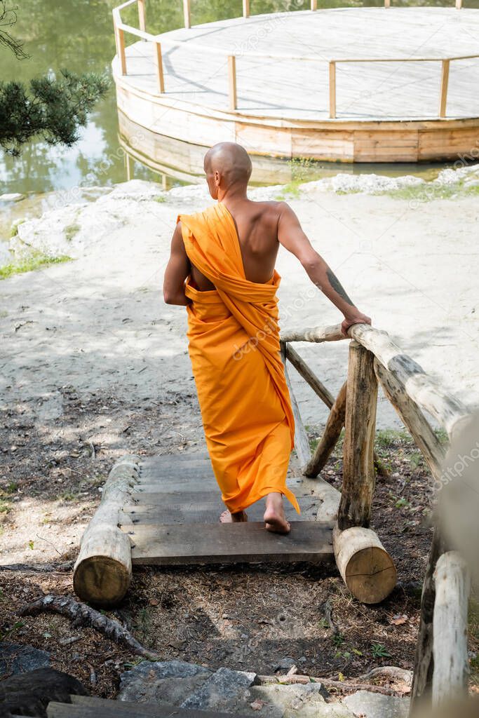 back view of hairless man in traditional buddhist clothes walking on wooden stairs in forest