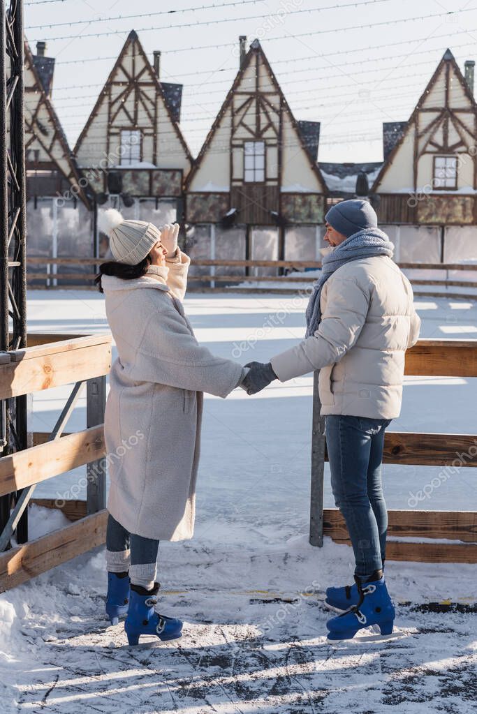 full length of happy young couple in winter hats and ice skates holding hands outside