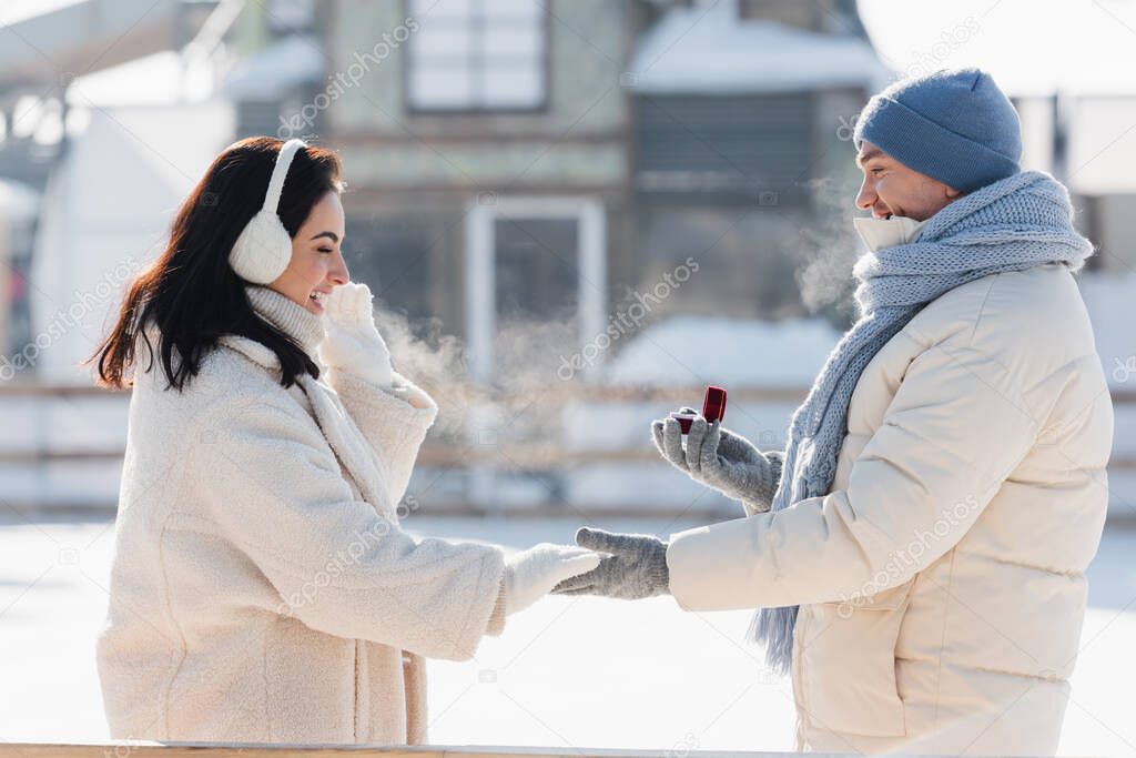 side view of man in winter hat holding box with wedding ring near happy young woman in ear muffs
