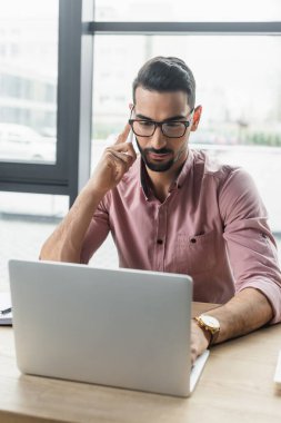 Gözlüklü Müslüman işadamı akıllı telefonla konuşuyor ve ofiste dizüstü bilgisayar kullanıyor. 