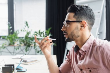 Gözlüklü Müslüman işadamının ofisteki akıllı telefondan sesli mesajı kaydedişinin yan görüntüsü 