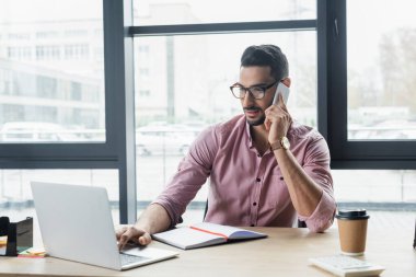 Müslüman işadamı akıllı telefondan konuşuyor ve bilgisayarı kahvenin yanında kullanıyor. 
