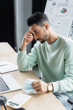 Ofiste laptopun yanında tıbbi maske tutan yorgun Müslüman işadamı 