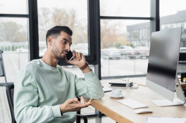 Müslüman işadamı bilgisayarın yanında akıllı telefonla konuşuyor ve ofiste kahve içiyor. 