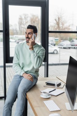 Gündelik giysiler içinde Müslüman bir işadamı telefon başında defterlerin yanında konuşuyor ve ofiste kahve içiyor. 