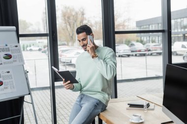 Ofiste akıllı telefonla konuşurken dizüstü bilgisayara bakan olumlu Arap işadamı 