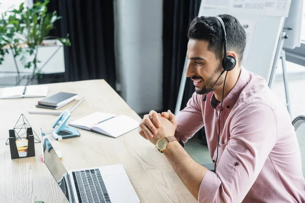 Side view of positive arabian businessman in headset having video call on laptop in office