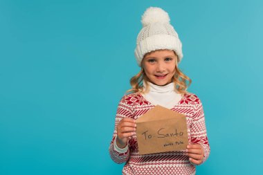Sıcak şapkalı ve kazaklı mutlu kız mavi üzerinde Noel Baba 'ya mektup gösteriyor.