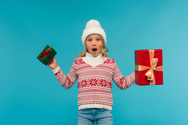 astonished girl in knitted sweater showing red and green gift boxes while looking at camera isolated on blue