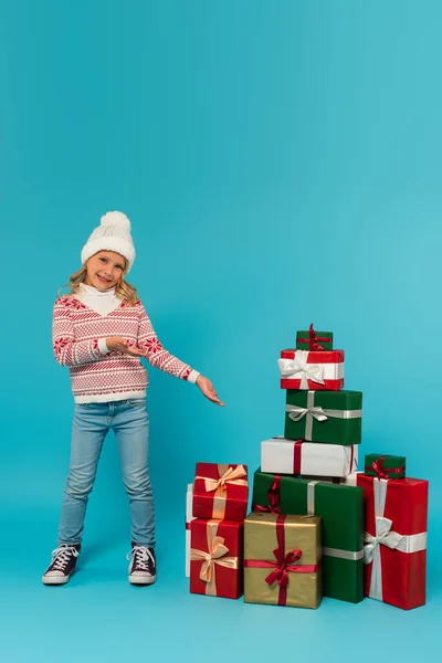 头戴针织帽子 毛衣和牛仔裤 面带微笑的女孩 指着一堆蓝色礼物 — 图库照片