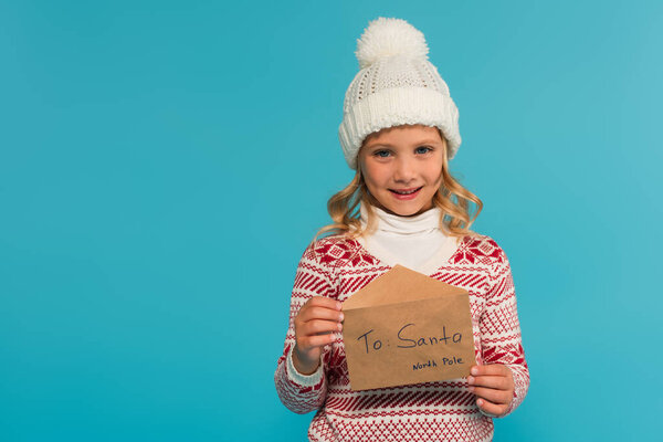 happy girl in warm hat and jumper showing letter to santa isolated on blue