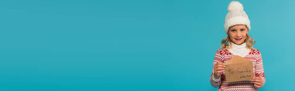 pleased girl in warm hat and jumper holding letter ro santa isolated on blue, banner