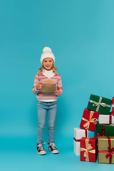 full length view of girl in warm sweater and jeans holding letter to santa near gift boxes on blue