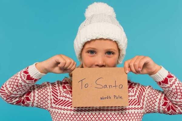 positive child in knitted hat holding letter to santa near face isolated on blue