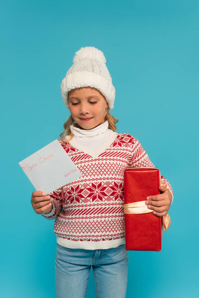 pleased child in warm sweater and hat holding present and letter to santa clause isolated on blue
