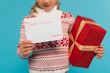 cropped view of blurred child with red gift box and letter to santa clause isolated on blue clipart