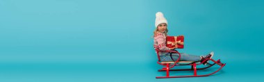 girl in warm hat and sweater sitting in sled with red gift box on blue, banner clipart
