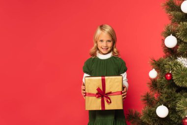 Yeşil elbiseli neşeli kız elinde süslü Noel ağacının yanında kırmızı renkte izole edilmiş bir hediye tutuyor.