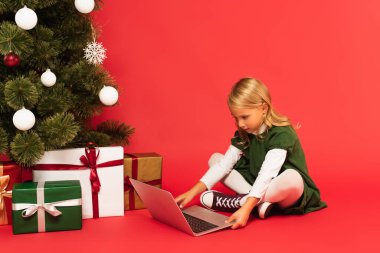 Yeşil elbiseli kız kırmızı üzerinde noel ağacı altında hediye kutularının yanında dizüstü bilgisayar kullanıyor.