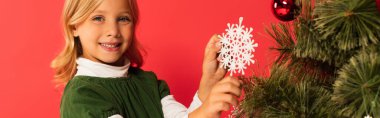 Kırmızı pankartta izole edilmiş Noel ağacını süslerken kameraya gülümseyen neşeli çocuk.