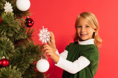 Mutlu çocuk elinde dekoratif kar taneleri tutuyor kırmızı üzerine izole edilmiş Noel ağacı yakınında.