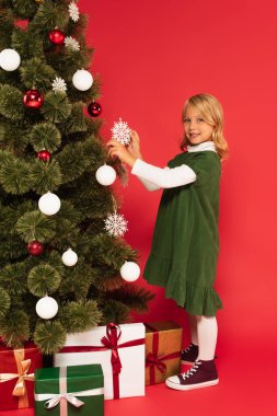 Yeşil elbiseli şık bir kız kameraya gülümserken Noel ağacını kırmızıyla süslüyor.