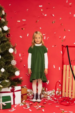 Yeşil elbiseli ve spor ayakkabılı mutlu kız kızağın yanındaki kameraya gülümsüyor ve kırmızı üzerinde noel ağacı altında hediyeler veriyor.