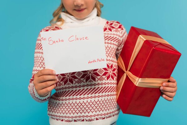 cropped view of blurred child with red gift box and letter to santa clause isolated on blue