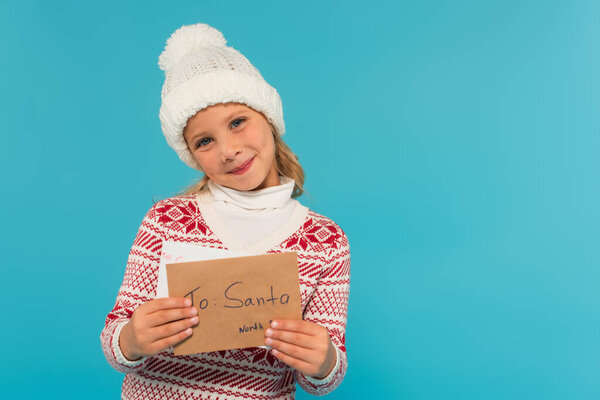 joyful child in knitted hat and sweater showing letter to santa isolated on blue
