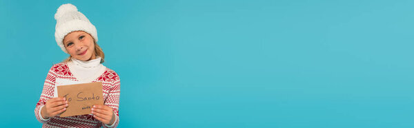 smiling girl in warm jumper and hat holding letter to santa isolated on blue, banner