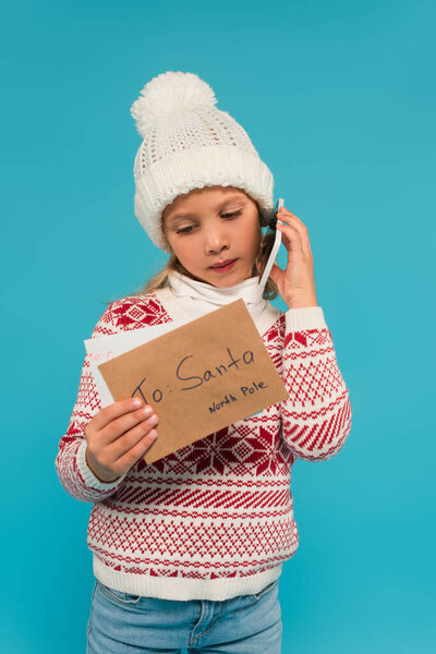 kid in knitted hat and jumper calling on smartphone while holding letter to santa isolated on blue
