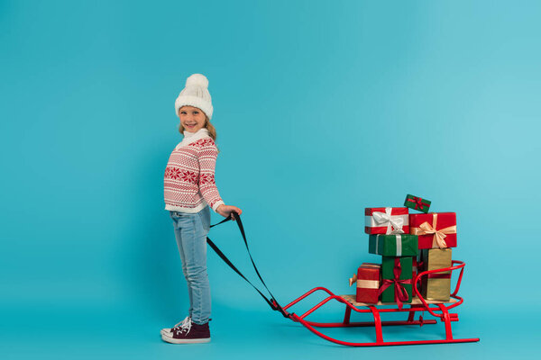 full length view of joyful girl in knitted sweater and hat near sled with presents on blue