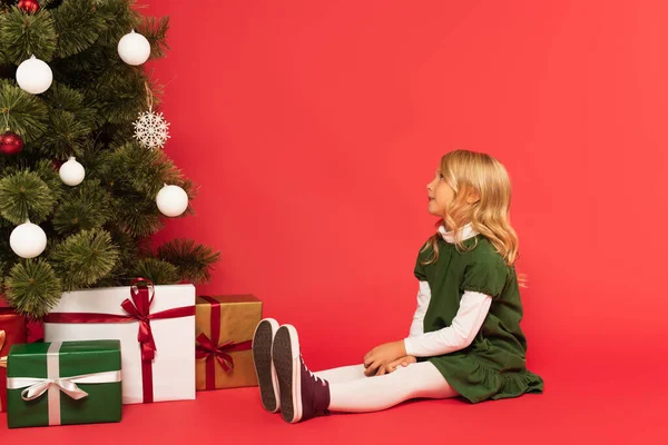身穿绿色衣服的女孩坐在红色礼品盒旁边 看着圣诞树 — 图库照片