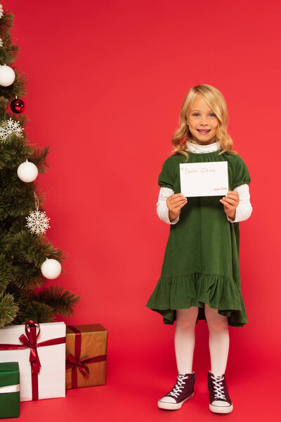 smiling girl with letter to santa clause near presents under christmas tree on red