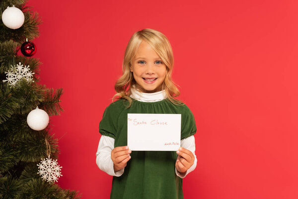 joyful girl holding letter to santa clause near decorated christmas tree isolated on red