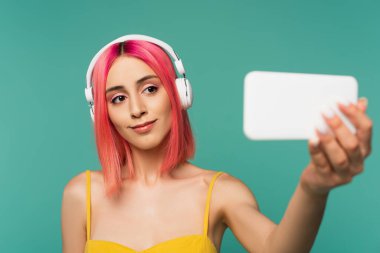 Pembe boyalı saçlı genç bir kadın kulaklıkla müzik dinlerken mavi renkte selfie çekiyor.