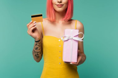 Pembe boyalı saçlı, elinde hediye paketi olan mutlu bir genç kadın ve mavi üzerine izole edilmiş bir kredi kartı. 