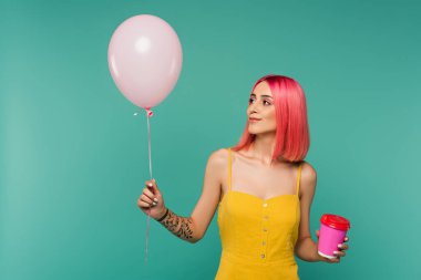 Pembe boyalı saçlı genç bir kadın elinde kağıt bardakla mavi renkli balona bakıyor. 