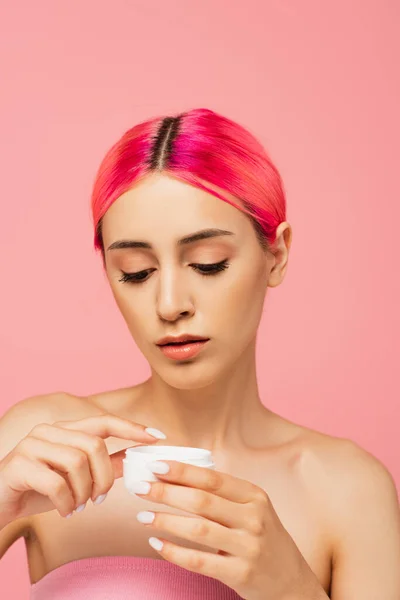染了头发的年轻女人看着用粉红隔离的化妆品霜的容器 — 图库照片
