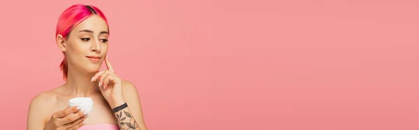 容器を持ちながらカラフルな髪の笑顔で若い女性を入れ墨し ピンク バナーに孤立した顔クリームを適用します — ストック写真