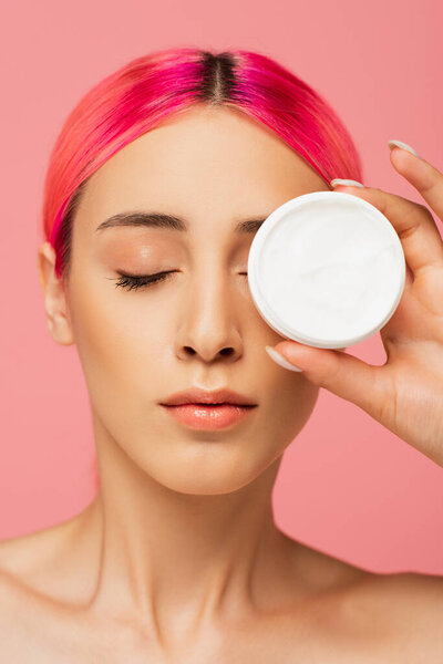 young woman with colorful hair covering eye while holding container with cosmetic cream isolated on pink