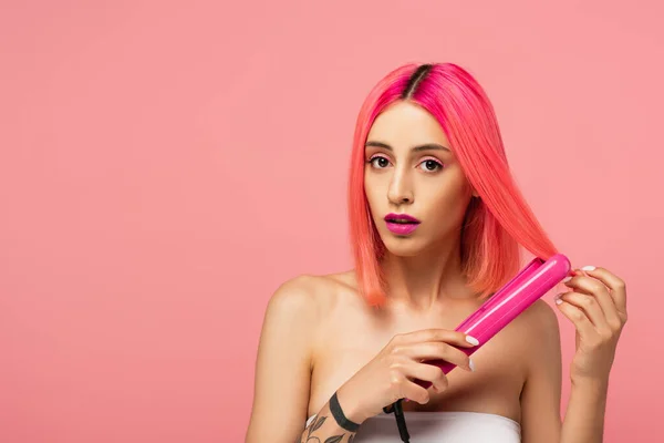 tattooed young woman with colorful hair using hair straightener isolated on pink