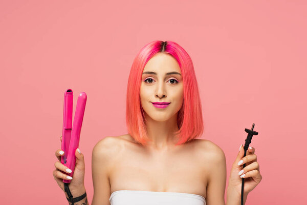 cheerful young woman with colorful hair holding hair straightener isolated on pink 