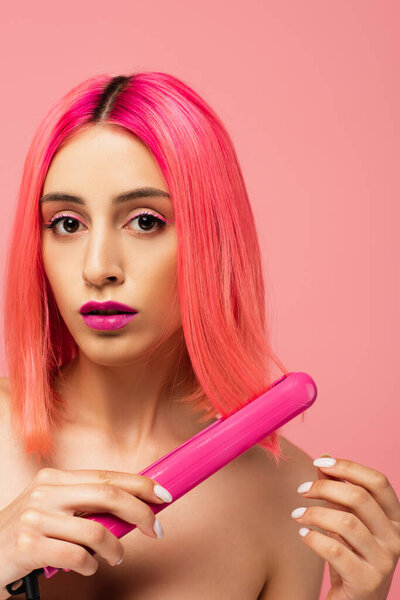 young woman with dyed hair using hair straightener isolated on pink 