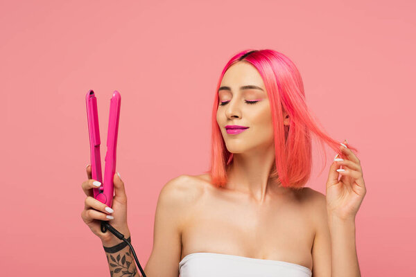 pleased young woman with colorful hair holding hair straightener isolated on pink 
