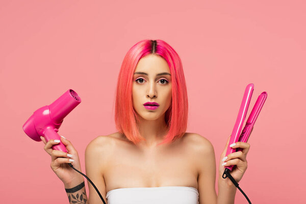 young woman with colorful hair holding hair dryer and straightener isolated on pink 